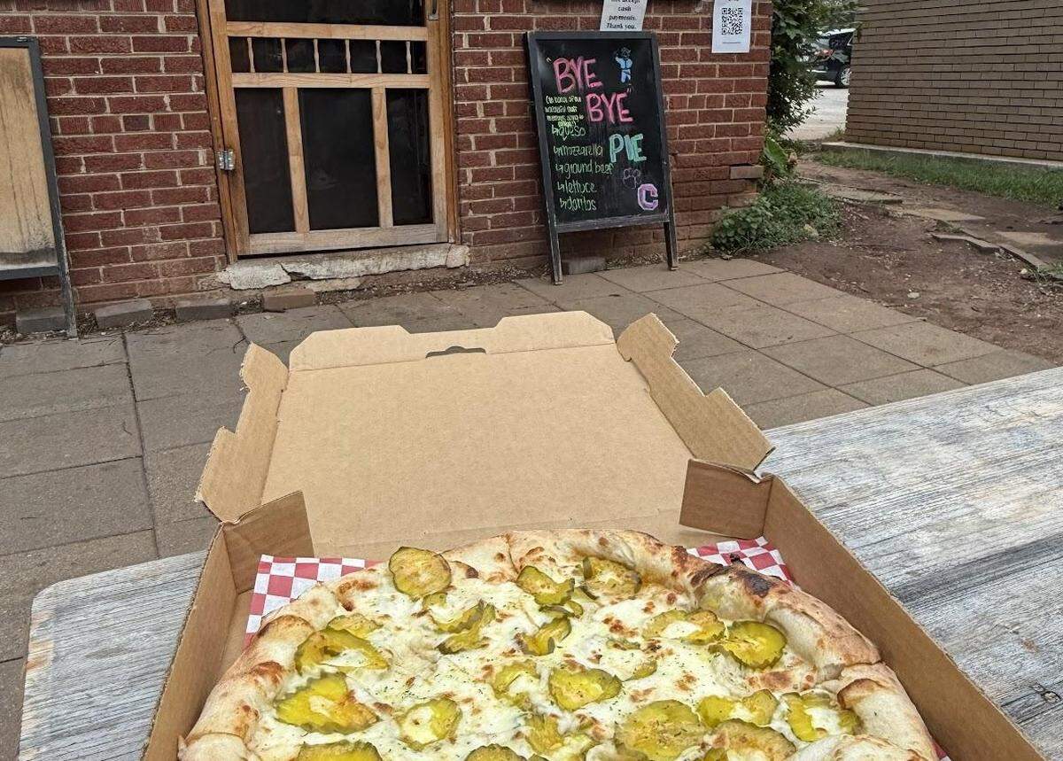 A first-person view of an open pizza box on a wooden picnic table. Inside is an uncut pizza with a white sauce base, topped with cheese and a large amount of sliced pickles. In the background is the brick entrance to the pizzeria with a chalkboard sign that reads “BYE BYE PIE.”