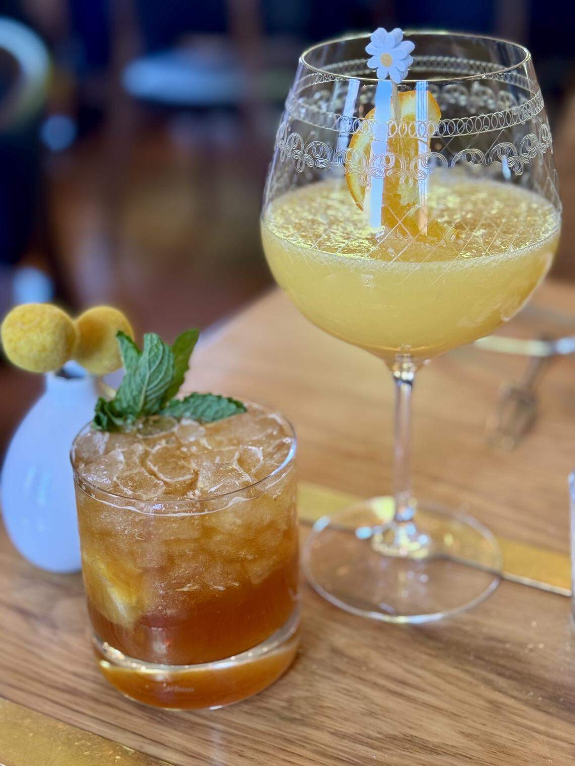 A beautifully composed shot of two craft cocktails on a light wood tabletop. In the background, a Zero Mimosa is served in a delicate, oversized stemmed glass with intricate etched patterns, garnished with an orange slice and a whimsical white daisy stirrer. In the foreground, a Pimm’s Cup sits in a classic rocks glass, packed with crushed ice and topped with a vibrant sprig of fresh mint. The drinks are presented alongside a small white vase with yellow billy ball flowers, capturing the elegant and refreshing aesthetic of a high-end brunch service.