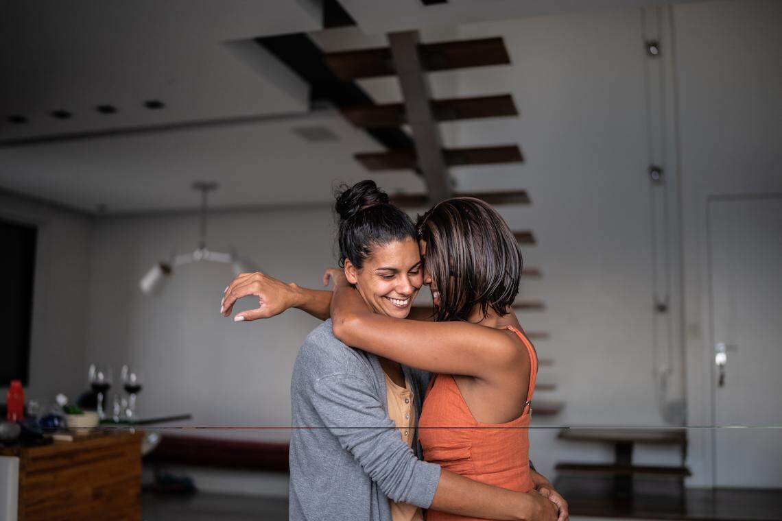 Two people stand and embrace each other affectionately in a modern home’s living area. One person, with their back mostly to the camera, has their arms wrapped around the other, who is facing forward and smiling with their eyes closed.
