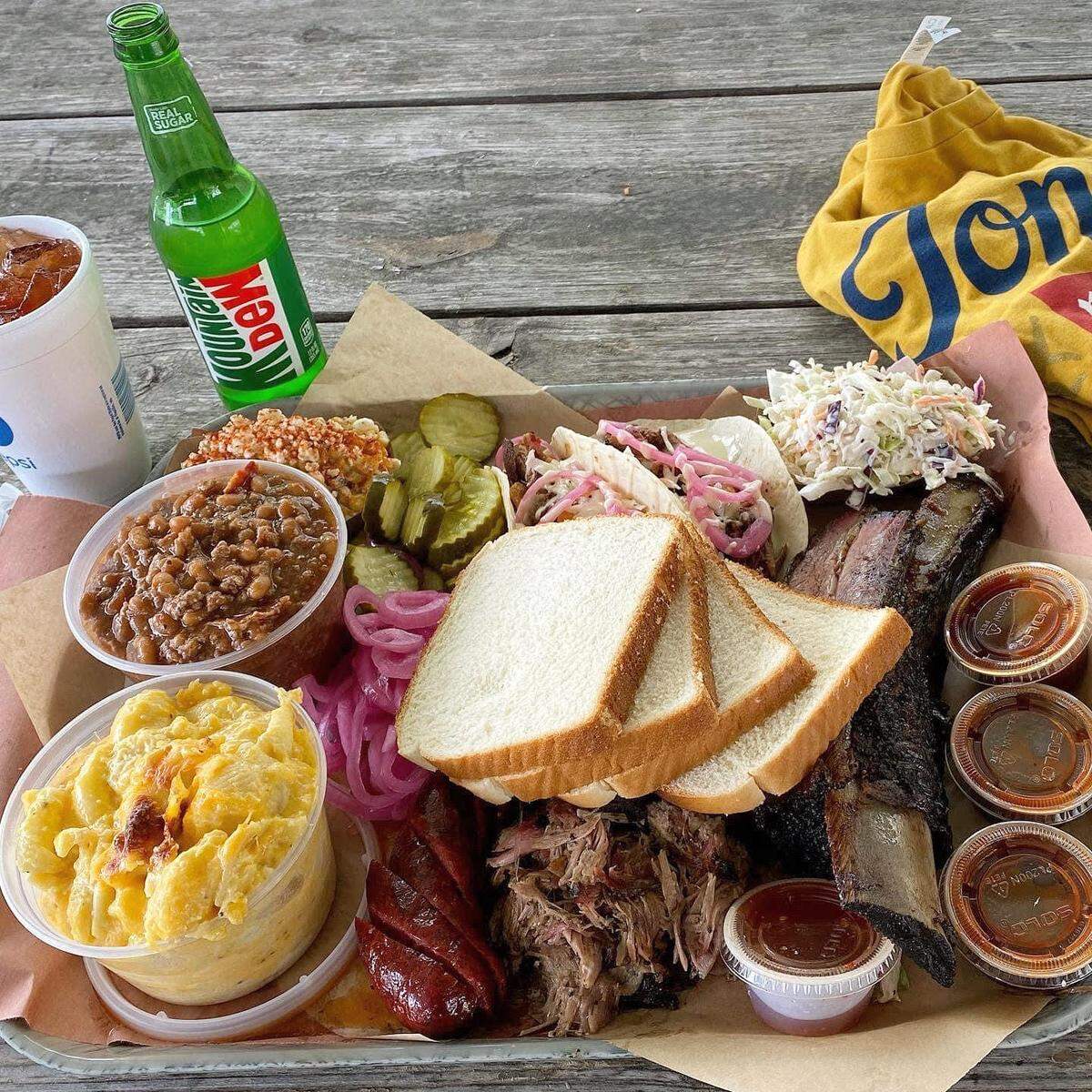 A generous platter of barbecue food is presented on a metal tray lined with brown butcher paper, set on a rustic wooden picnic table. The tray holds a variety of meats and sides: several large, dark beef ribs, a pile of pulled pork, and slices of sausage. Sides include a clear plastic container of creamy macaroni and cheese, a bowl of baked beans, a portion of red rice, slices of dill pickles, and pickled red onions. Three slices of white bread are stacked in the center. Four small plastic cups with various barbecue sauces are also on the tray. To the left, a white cup with a lid and straw holds a drink, and a green glass bottle of Mountain Dew is visible. A yellow t-shirt with “Jon G’s” embroidered in blue is partially visible in the upper right corner.