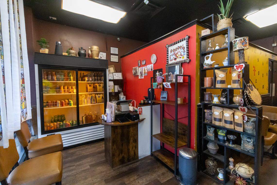 A view of the restaurant’s entrance and counter area. A vibrant red accent wall serves as a backdrop for the wooden host stand. To the left, a large refrigerated display case is filled with beverages. To the right, a black wooden shelving unit acts as a mini-market, displaying bags of Ethiopian coffee beans, traditional pottery, and cultural artifacts for guests to browse or purchase.