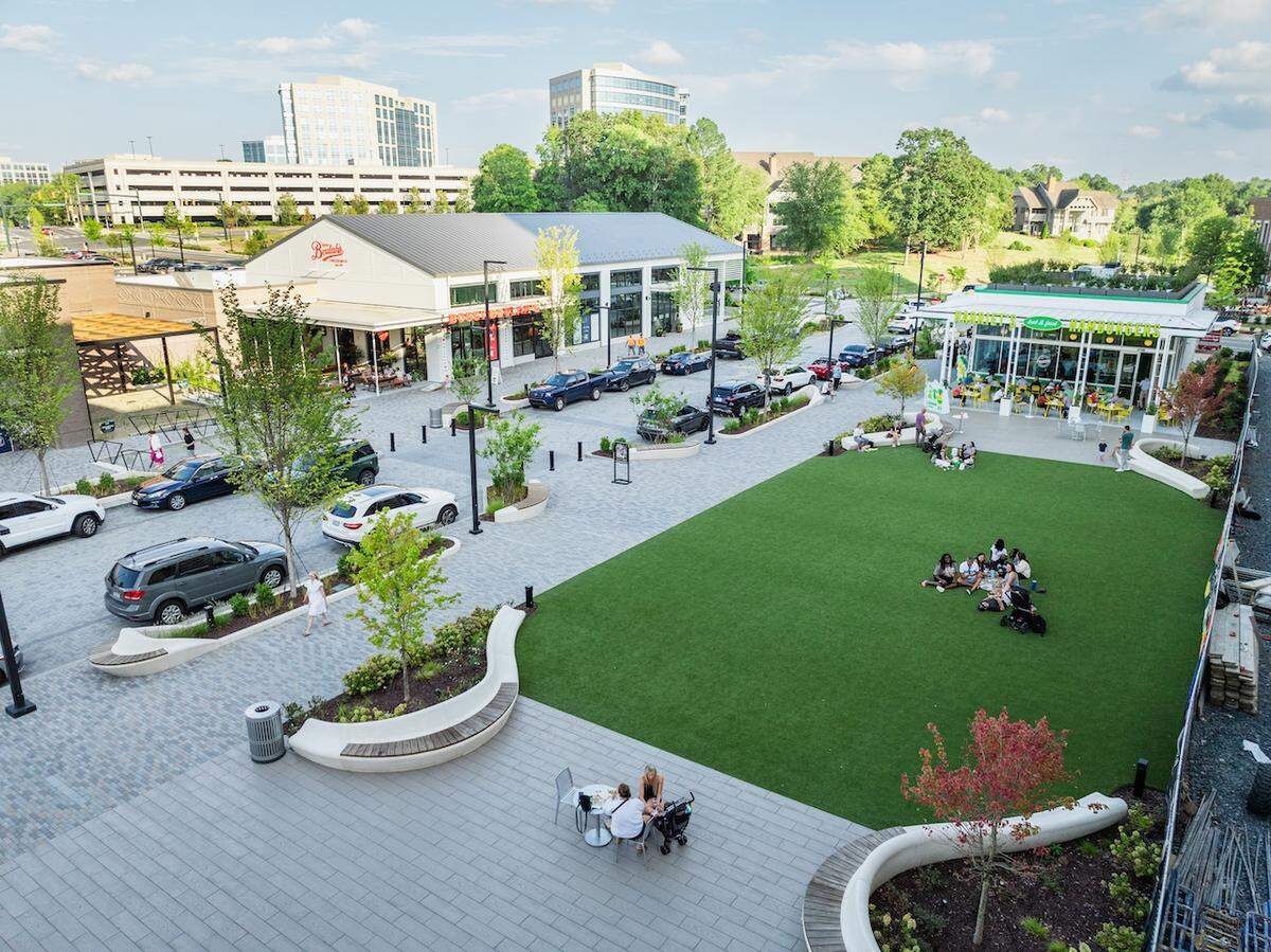 A high-angle, aerial shot of a modern mixed-use development plaza on a sunny day. The scene features a large, manicured green lawn where groups of people are sitting, surrounded by gray paver pathways, curved concrete seating, and modern white commercial buildings.