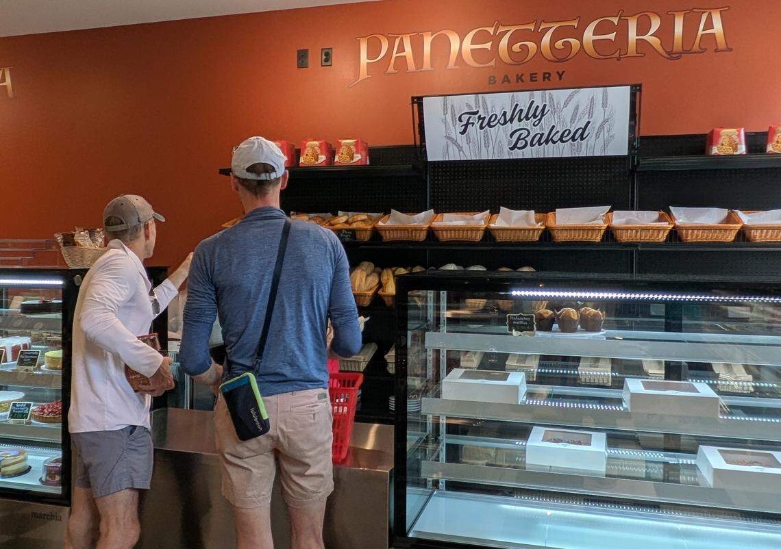Two customers browse the “Panetteria” bakery section. In the background, a sign reads “Freshly Baked” above shelves of bread loaves. In the foreground, glass display cases show an assortment of cakes, muffins, and pastries.