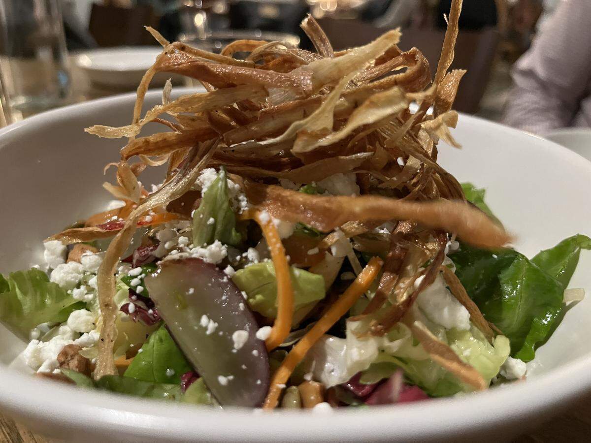 A close-up shot of a white bowl filled with a fresh salad. The base consists of bright green lettuce, red radicchio, and shredded carrots. The salad is mixed with crumbled white cheese (feta or goat cheese) and halved purple grapes. It is heavily topped with a tall mound of thin, crispy, golden-brown fried strips, possibly of parsnip or potato. The setting is dimly lit, suggesting a restaurant interior, with a wooden table visible underneath the bowl.
