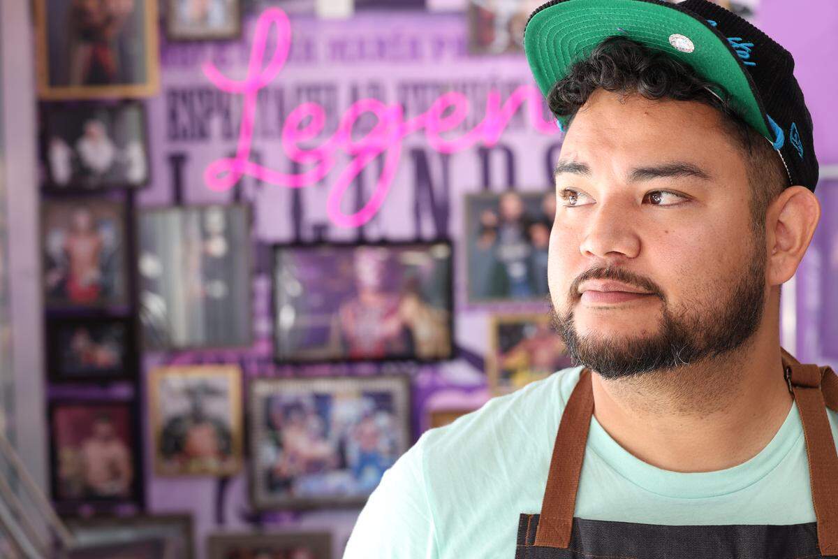 A tight, close-up shot of an ice cream shop owner wearing a mint-colored t-shirt, apron, and a backward green baseball cap, looking off to the right. The background is blurred, revealing a purple wall with framed photos and a bright pink neon sign.