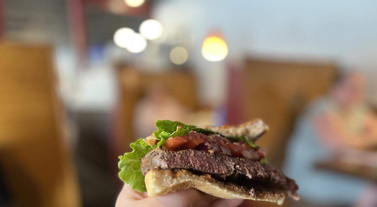 A macro shot of a steak sandwich being held. The focal point is the thick slice of grilled steak, cooked to a pink medium-rare. Above the steak are layers of bright green lettuce and a fresh topping of diced red tomatoes and onions, all on a crusty, toasted roll. The background is a blurry restaurant interior.