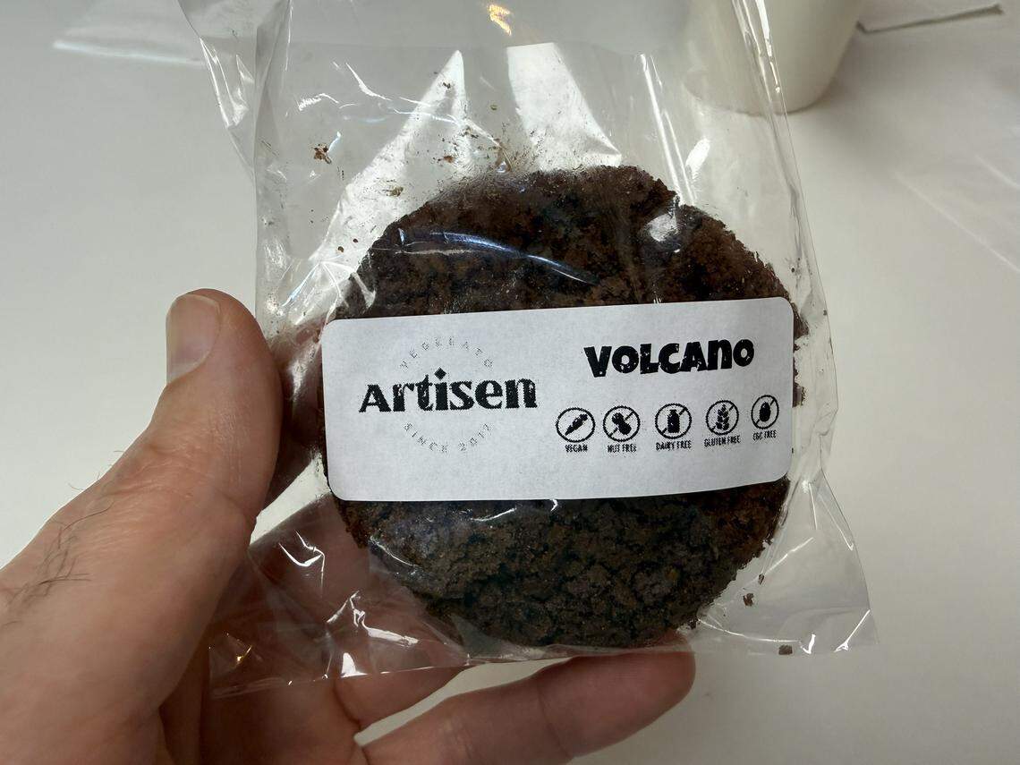 A close-up shot of a hand holding a large, dark chocolate cookie sealed in a clear plastic bag. The white label identifies it as a “VOLCANO” cookie from Artisen. The label also features icons confirming the cookie is vegan, nut-free, dairy-free, gluten-free, and egg-free, emphasizing the shop’s focus on inclusive treats.
