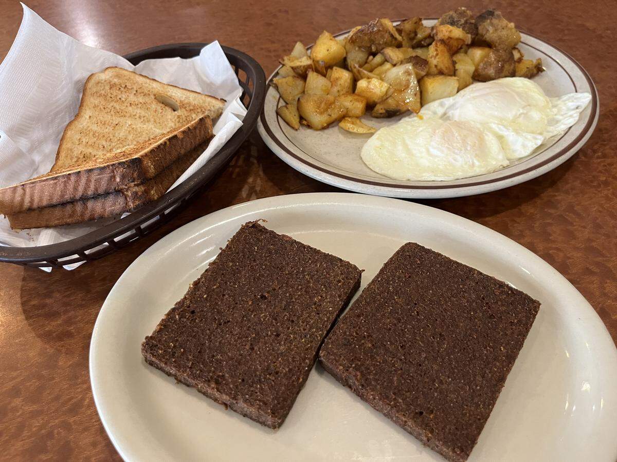 A breakfast plate featuring two fried eggs and a pile of home fried potatoes and onions, alongside a separate plate holding two thick, rectangular slices of dark liver mush. A basket of toasted white bread is also visible on the table.