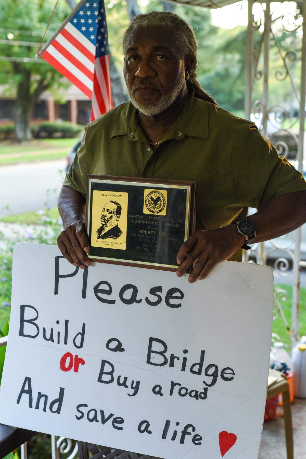 Robert Neal, 69, holds a sign reading “Please Build a Bridge or Buy a road And save a life” outside his home in the historically Black Logan neighborhood in Concord, N.C., on Friday, Aug. 25. 2023. The Lincoln Street Bridge, blocks away from the city-owned unit where three children died in a fire, has been closed for more than a year.