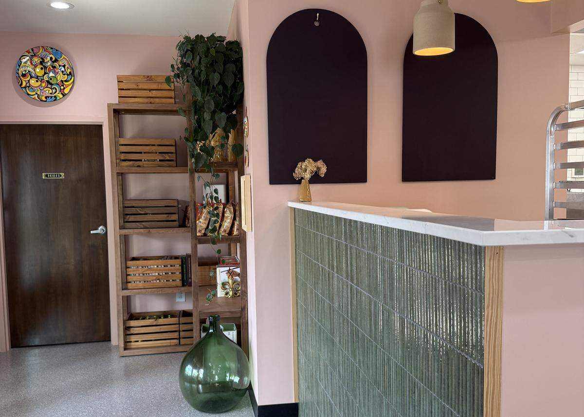 The interior of a shop with soft pink walls, a green tiled counter with a marble top, and a tall wooden shelf holding crates and bottles.