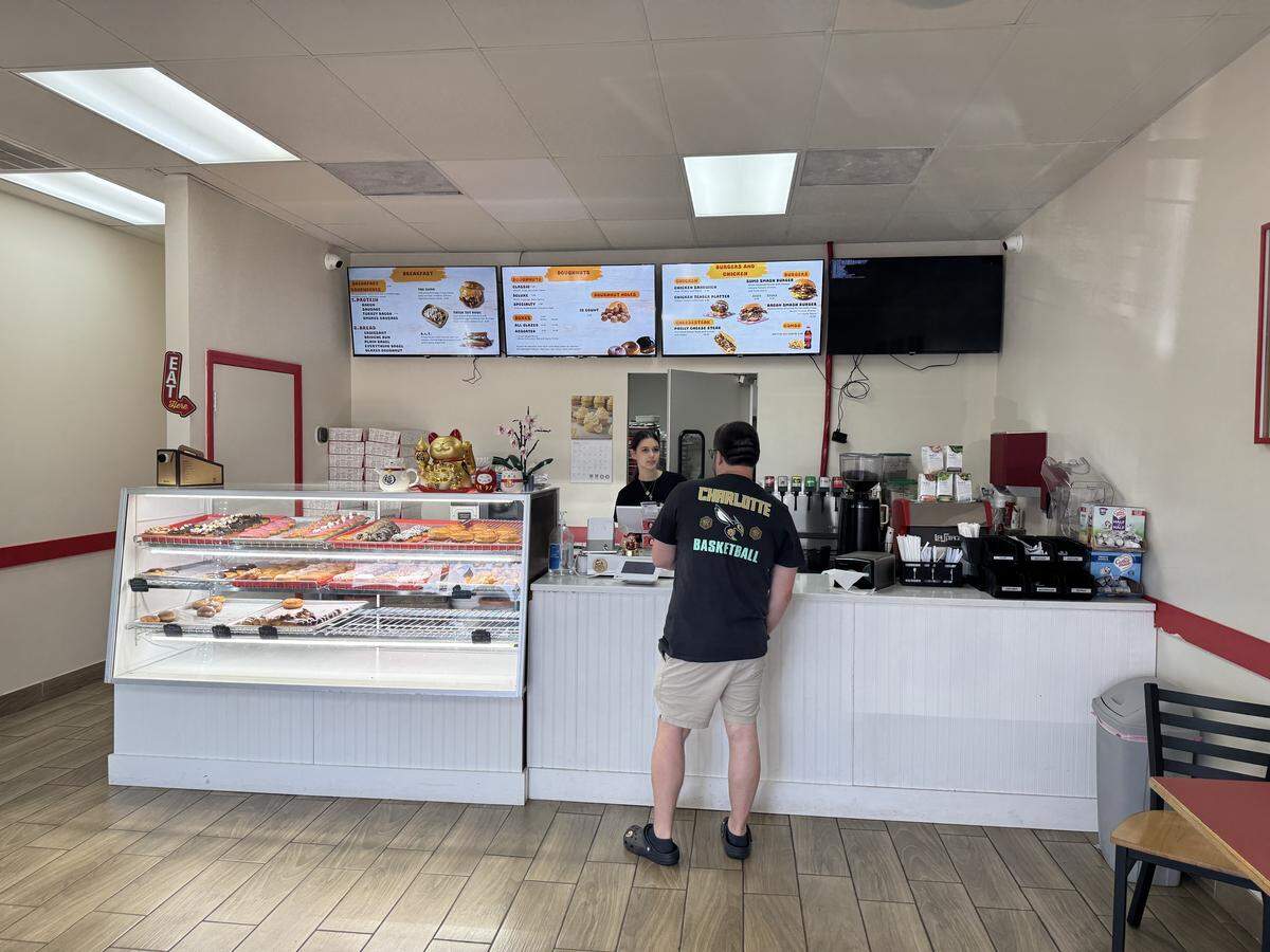 The interior of the doughnut shop. A glass display case on the left is filled with several rows of colorful, frosted doughnuts. Behind a white wooden counter, a staff member is assisting a customer wearing a “Charlotte Basketball” t-shirt. Three digital menu boards hang above the counter, displaying breakfast items, doughnuts, and burgers. A golden “lucky cat” (Maneki-neko) figurine sits on top of the display case.