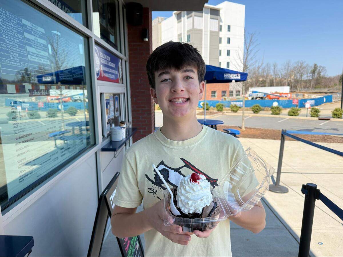 A teenager smiles outside an ice cream shop while holding a chocolate sundae in a clear plastic container.