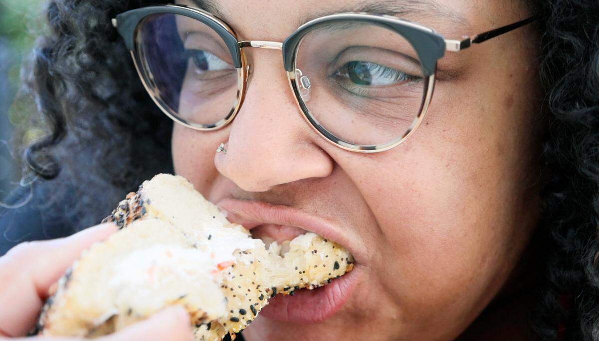 An intimate, close-up photo shows a reporter with glasses and a nose piercing in the middle of eating an everything bagel. Her mouth is open around the bagel, which is heavily coated in seeds and spread with a thick layer of cream cheese.