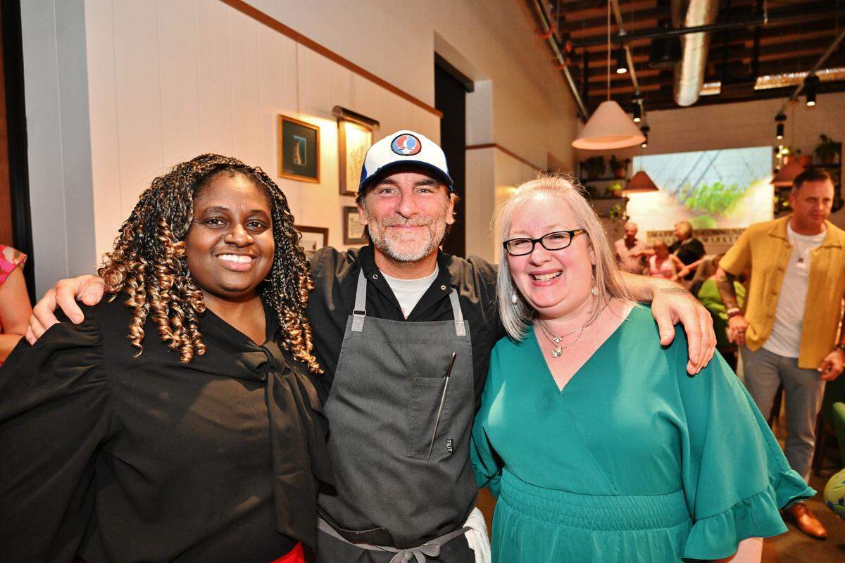 A photograph of three people standing together, smiling, with their arms around each other in a warmly lit, modern, casual interior space. From left to right: a person with dark, curly hair wearing a black blouse; a chef in the center wearing a dark apron over a white shirt and a white and blue baseball cap; and a person with short gray hair and glasses wearing a teal green dress. The background shows white walls with framed artwork, exposed ductwork on the ceiling, pendant lighting, and other people in the distance.