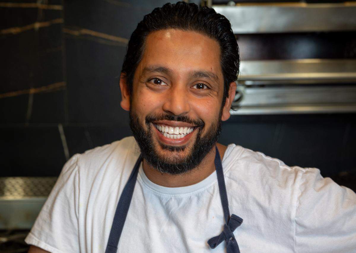 A close-up, high-resolution portrait of a smiling chef with a well-groomed dark beard and slicked-back dark hair, wearing a simple white t-shirt and a dark apron. The background is slightly out of focus, showing a dark, marbled wall and the metallic surfaces of a commercial kitchen.