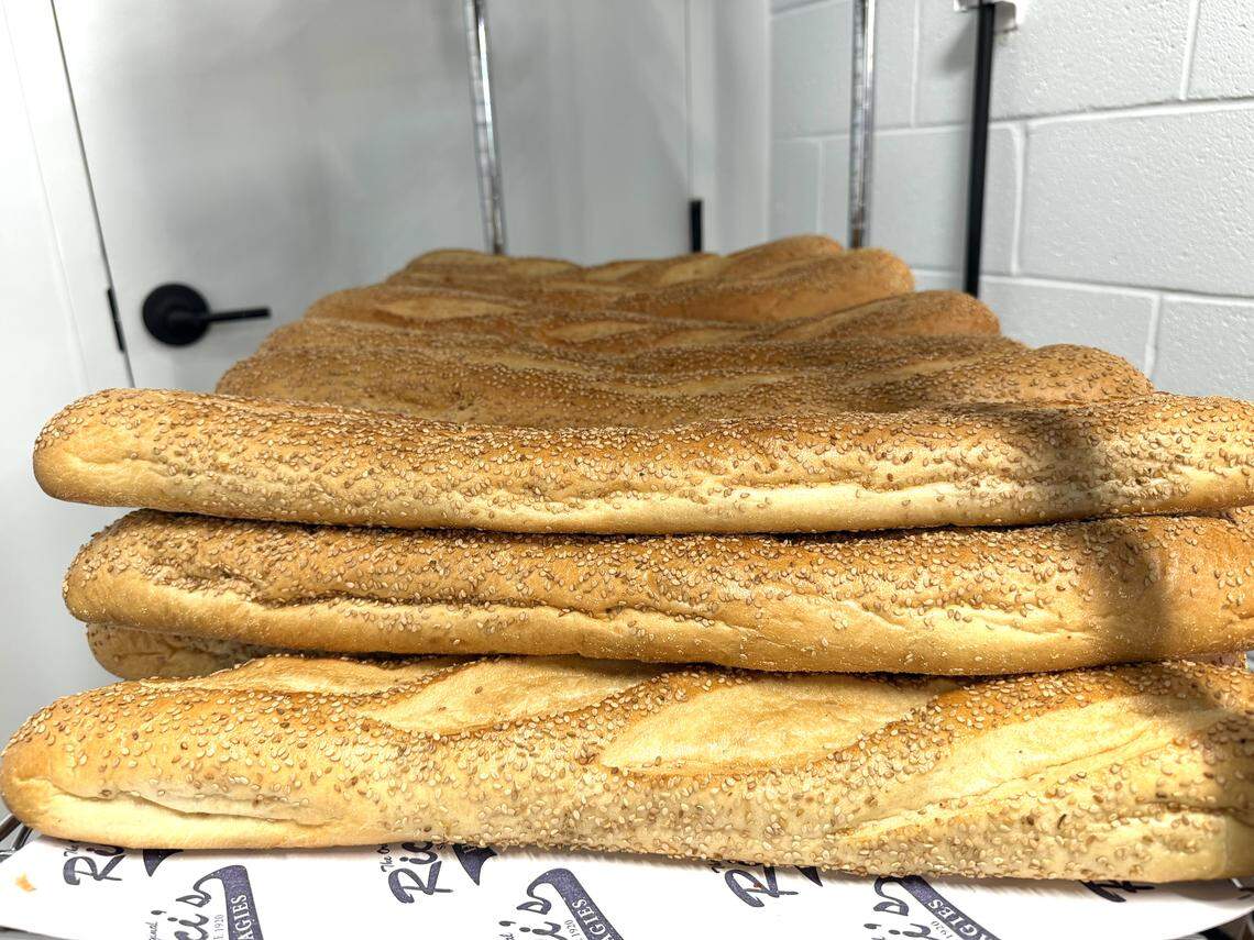 A close-up shot from the kitchen area shows a stack of fresh, artisanal bread with several long, golden-brown baguettes are heavily coated in sesame seeds.