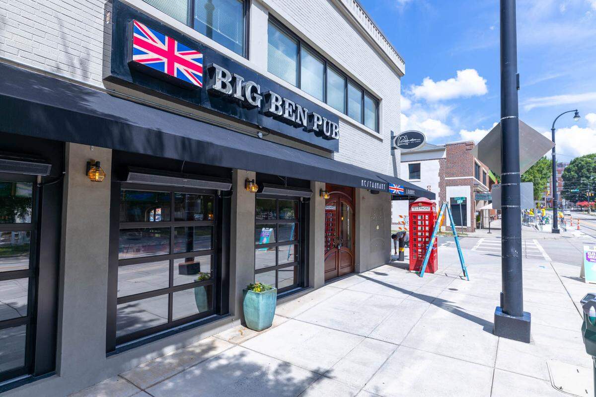 A wide, eye-level shot of the “Big Ben Pub” exterior on a sunny day. The restaurant, located in a light-colored brick building, has a large black awning with the pub’s name and a British flag. A classic red telephone booth stands on the sidewalk next to the entrance, and the street and a blue sky with clouds are visible in the background.