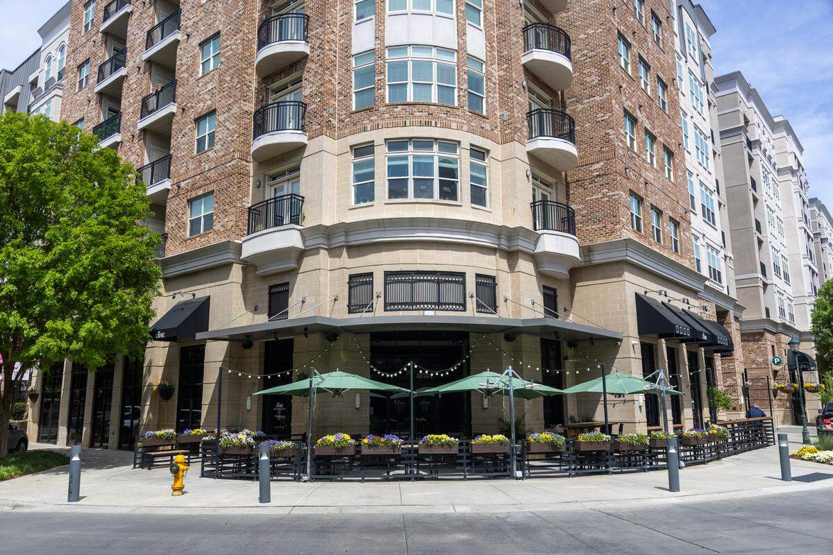 A wide exterior shot of The Craic, located on the ground floor of a multi-story brick and stone apartment building. The restaurant occupies a prominent corner with a wraparound patio enclosed by a black metal fence and topped with green umbrellas. Black awnings with the pub’s logo hang over the large windows, and colorful flower boxes add a bright touch to the sidewalk.