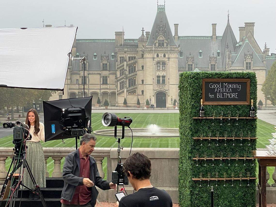 A behind-the-scenes photo of a TV shoot at the Biltmore Estate. With the iconic chateau in the background, a production crew films a host. A large prop hedge wall is set up as a champagne holder and features a chalkboard sign welcoming viewers from “Good Morning America.”