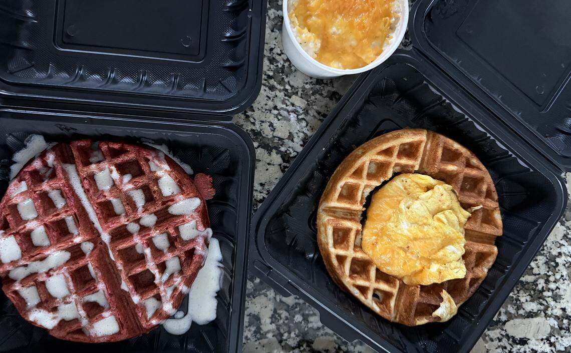 An overhead view of three black takeout containers on a granite countertop. The container on the left holds a vibrant red velvet waffle drizzled with white cream cheese icing. The container on the right contains a golden-brown Belgian waffle topped with a folded cheesy omelet in the center. At the top, a small white cup is filled with creamy grits topped with melted cheddar cheese.
