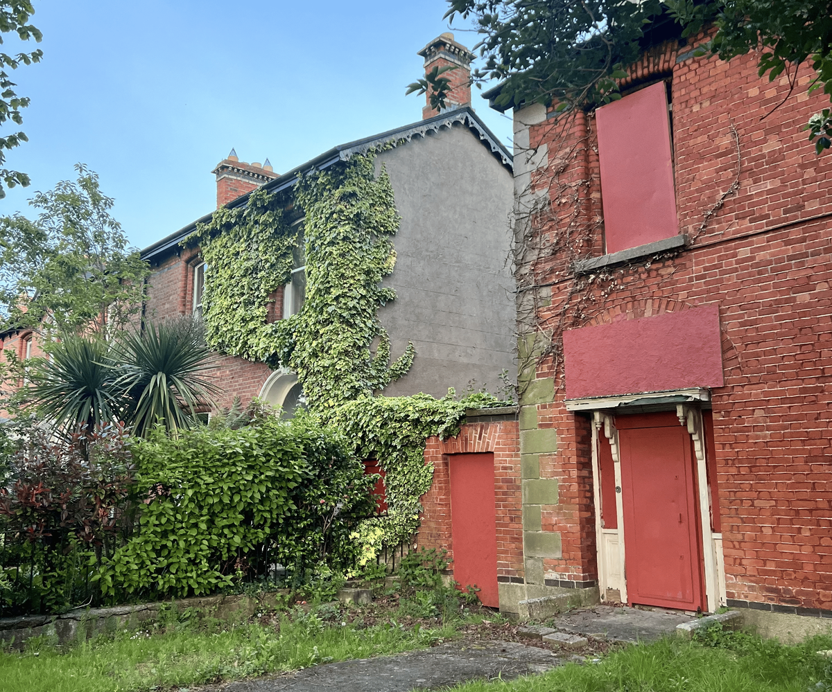 A view of the side of two old, red brick buildings, possibly row houses or townhomes. The buildings have overgrown bushes and green ivy climbing up the walls. The windows and a door on the building in the foreground are boarded up with red wooden panels. A small wooden porch with a white door and red panels is visible. The sky is a clear blue.