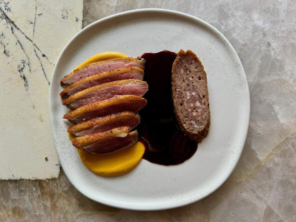 A beautifully plated gourmet dish is presented on a minimalist white plate with a subtle speckled rim, set on a light marble surface. On the left, five slices of perfectly cooked duck breast, showing pink meat and crispy golden skin, are fanned out over a vibrant yellow puree or sauce. To the right, a thick, oval slice of what appears to be a sausage or forcemeat is nestled in a rich, dark brown sauce or reduction that pools on the plate. A piece of textured white paper with dark lines is visible to the left of the plate.