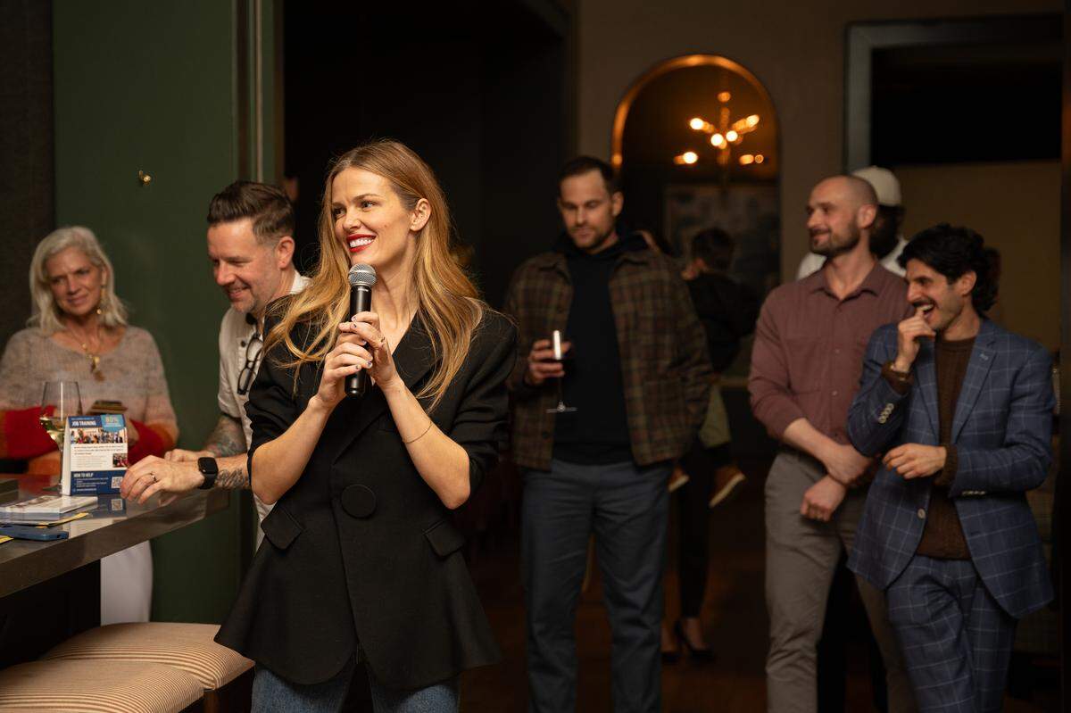 A person with long blonde hair and red lipstick wears a black blazer and speaks into a microphone at an indoor event. Several others are visible in the background, including one holding a glass of red wine.