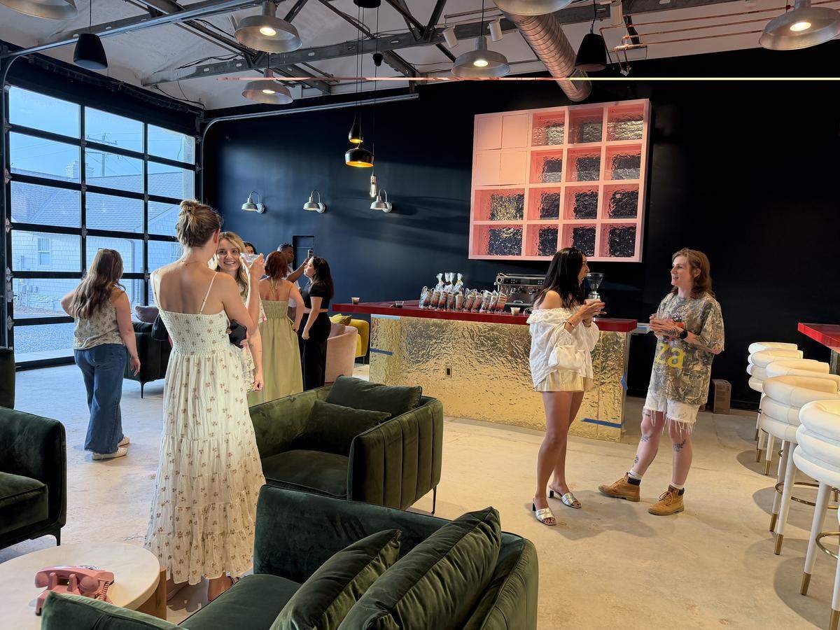 A wide shot of a modern, industrial-style lounge filled with people socializing. The space features high ceilings with exposed ductwork, dark matte walls, and a large glass garage-door window. Plush green velvet armchairs and sofas are scattered throughout. In the background, a gold-fronted bar with a red countertop sits under a large pink grid shelving unit.