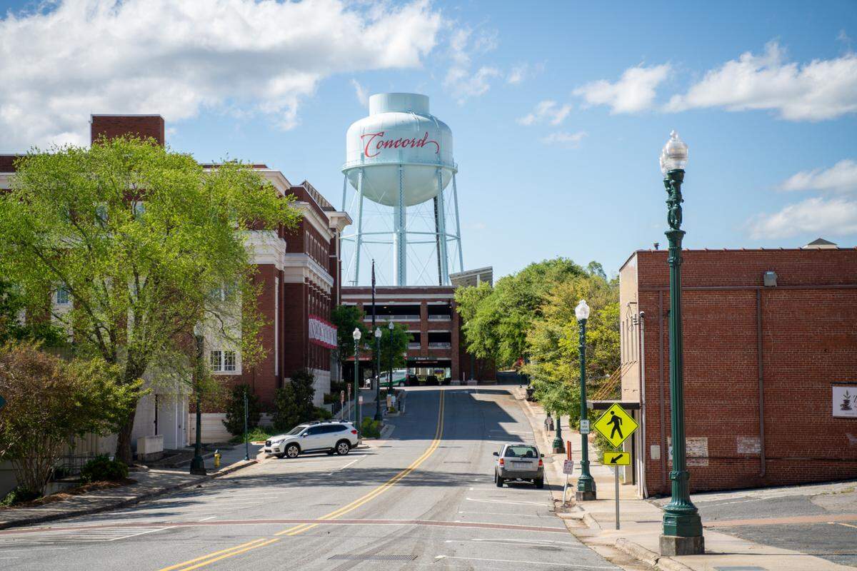 A wide, eye-level shot of a street in Concord, North Carolina. The asphalt road slopes uphill toward the background. At the top of the hill, a prominent light-blue water tower with the word “Concord” in red script is centered in the frame. The scene is flanked by brick buildings and vibrant green trees on a bright day with a blue sky and scattered clouds. A green lamppost and a pedestrian crossing sign are in the right foreground.