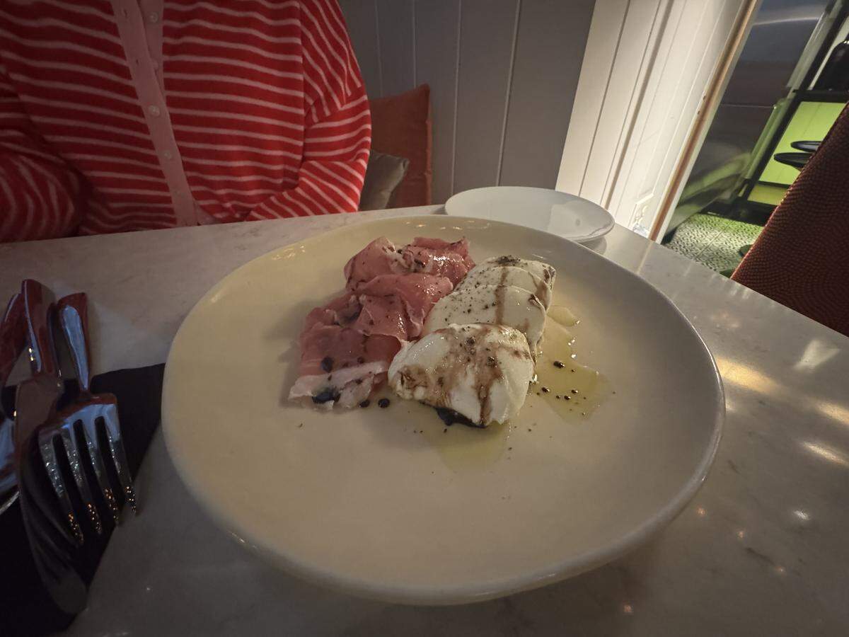 A close-up shot of an appetizer plate on a marble table. The off-white ceramic dish holds thin slices of pink prosciutto next to thick slices of fresh white mozzarella cheese. The cheese is drizzled with olive oil, a dark balsamic reduction, and a sprinkle of cracked black pepper. A person wearing a red-and-white striped shirt is partially visible in the background.