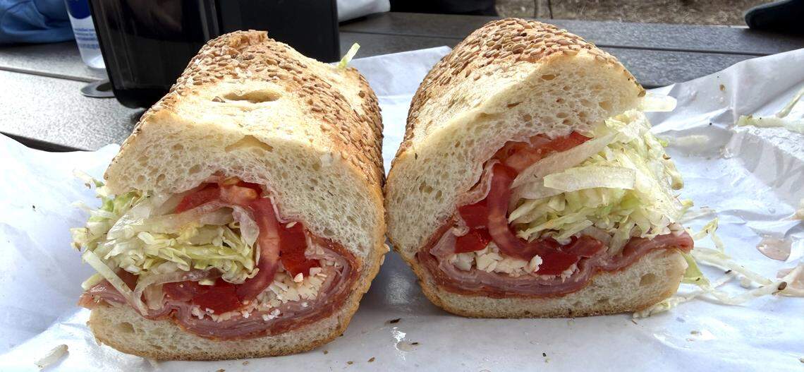 A close-up view of a large hoagie cut into two halves, served on white deli paper. The sandwich is made on a crusty long roll topped with sesame seeds and is packed with layers of sliced deli meats, red peppers, shredded lettuce, and white crumbled cheese.