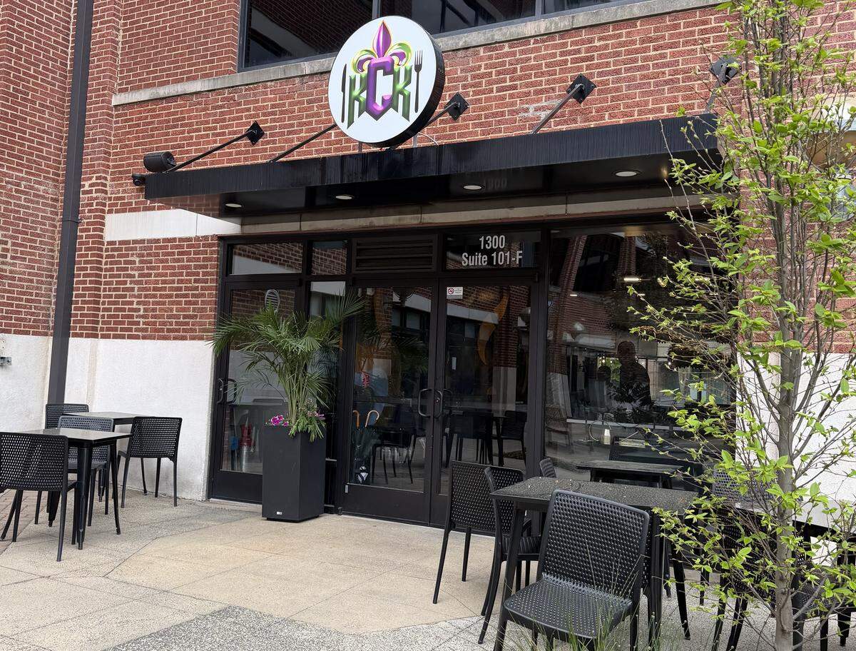 A set of black-framed glass double doors serves as a main entrance, with the address “1300 Suite 101-F” visible above the frame. A circular, backlit sign hangs above the black awning. It features a stylized “KCK” logo in green and white, topped with a purple and gold fleur-de-lis, flanking a knife and fork, hinting at a Cajun or Creole-influenced menu.