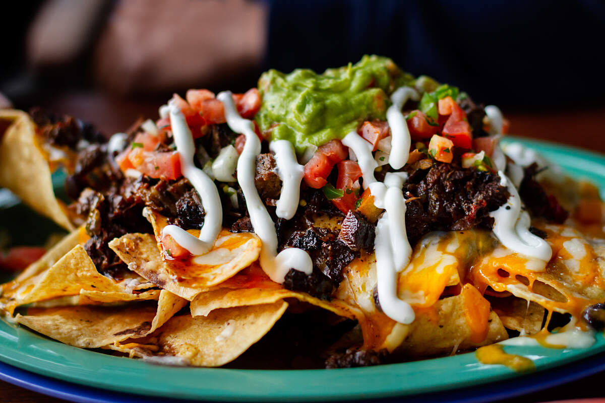 A close-up shot of a large plate of loaded beef nachos topped with melted cheese, shredded meat, diced tomatoes, sour cream, and guacamole.