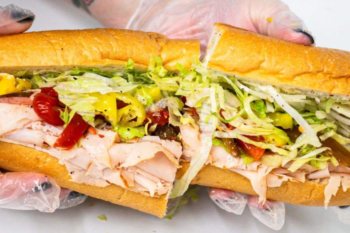 A close-up shot of a large hoagie sandwich, cut in the middle and held by hands in clear disposable gloves. The sandwich is filled with layers of thinly sliced deli meat, shredded lettuce, and a mix of red and yellow peppers on a crusty roll.