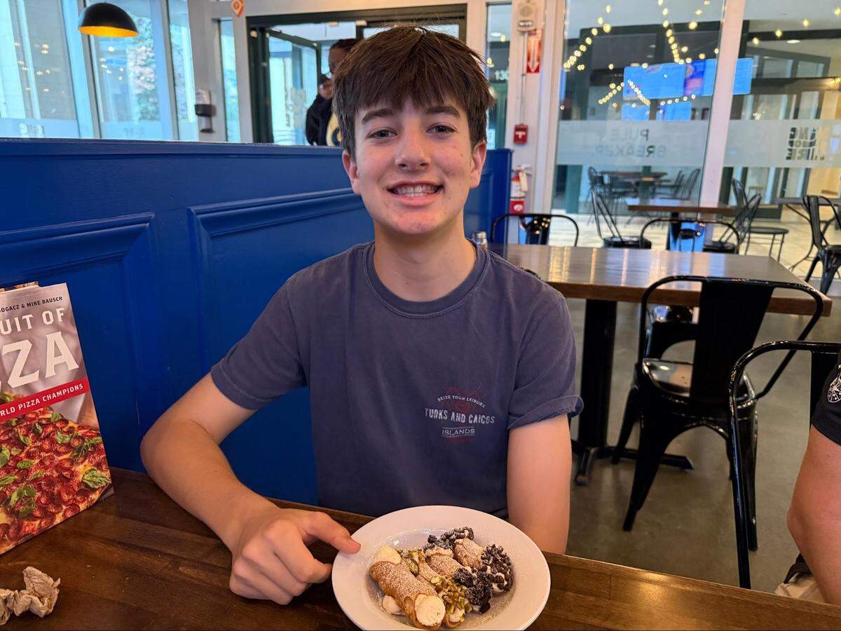 A young person in a blue “Turks and Caicos” t-shirt sits at a wooden restaurant table, smiling at the camera. In front of him is a plate of four mini cannolis garnished with chocolate chips, pistachios, and powdered sugar. A cookbook titled The Pursuit of Pizza is visible on the left.