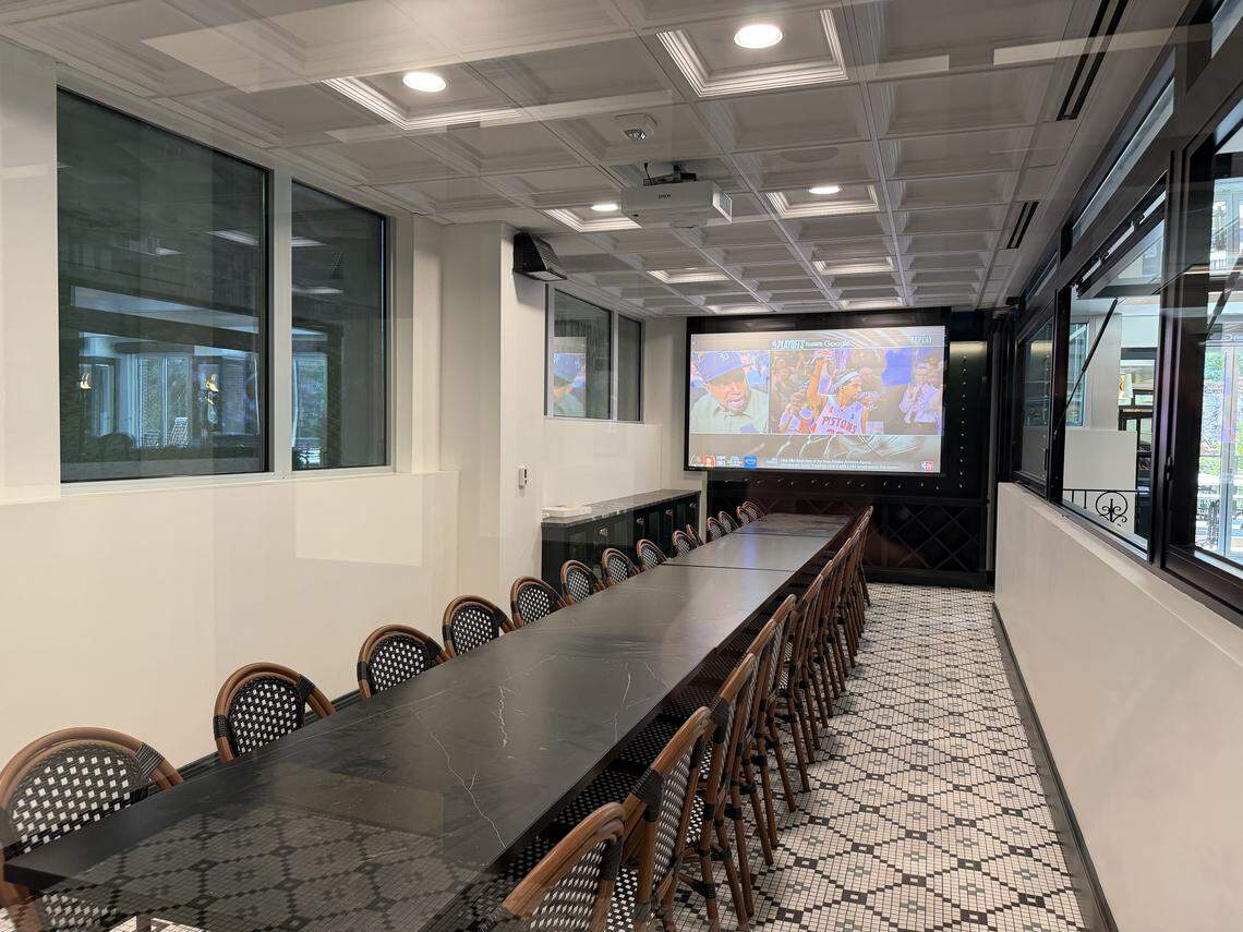 A long, dark wood communal table in a private dining or meeting room. A projector screen at the end of the room shows a sports broadcast.