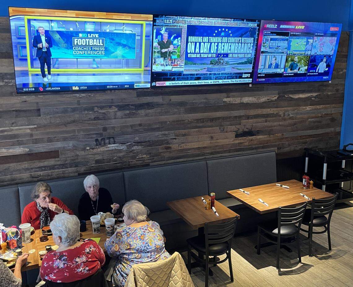An eye-level, medium shot showing four customers seated at a booth in a restaurant, eating and talking. They are sitting against a wall with horizontal wood paneling, and above them, three large televisions are mounted, all displaying live programming.