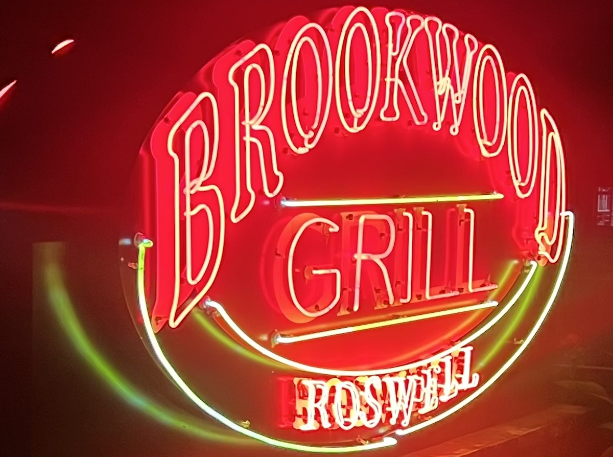 A glowing neon sign for “Brookwood Grill” in Roswell. The sign is circular with bright red neon lettering for “BROOKWOOD GRILL” and smaller white neon lettering at the bottom for “ROSWELL”. The sign is accented with curved yellow and green neon lines against a dark background.