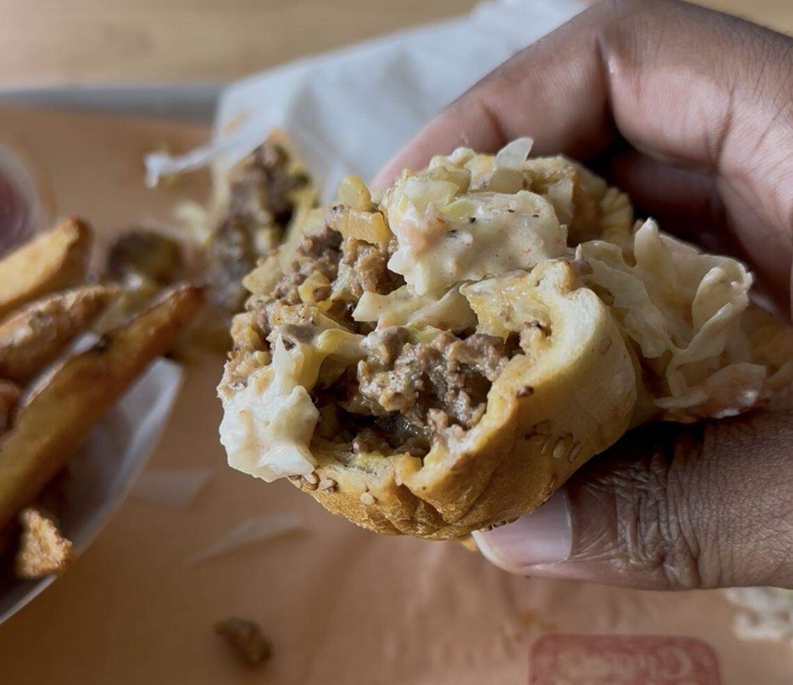 A close-up, first-person view of a hand holding a half-eaten sandwich filled with ground meat and coleslaw in a seeded bun. A side of thick-cut potato wedges is visible in the blurred background.