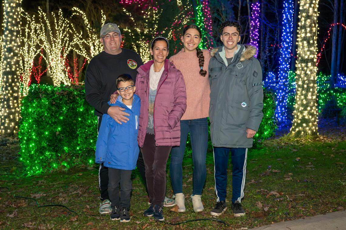 A family of four—a mother, father, teenage daughter, and young son — standing together outside at night, smiling. They are posing in front of a brilliant holiday light display where tree trunks and bushes are heavily wrapped in white, green, and multicolored string lights.