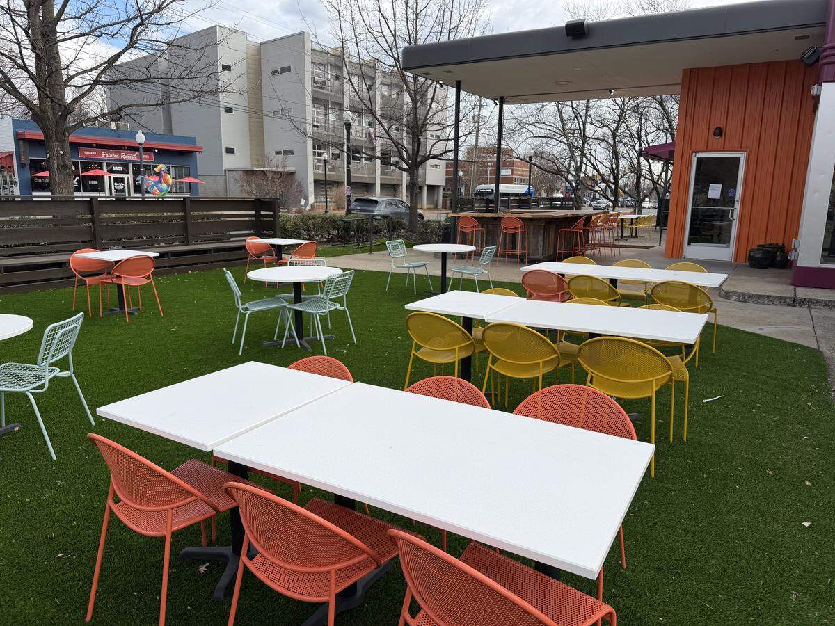 A view of a spacious outdoor dining patio at The Improper Pig, featuring artificial green turf and a variety of colorful metal mesh chairs in orange, yellow, and light blue. Several white rectangular and round tables are spread across the turf. In the background, an outdoor wooden bar sits under a covered area next to an orange corrugated metal wall, with a multi-story residential building and bare trees visible under an overcast sky.