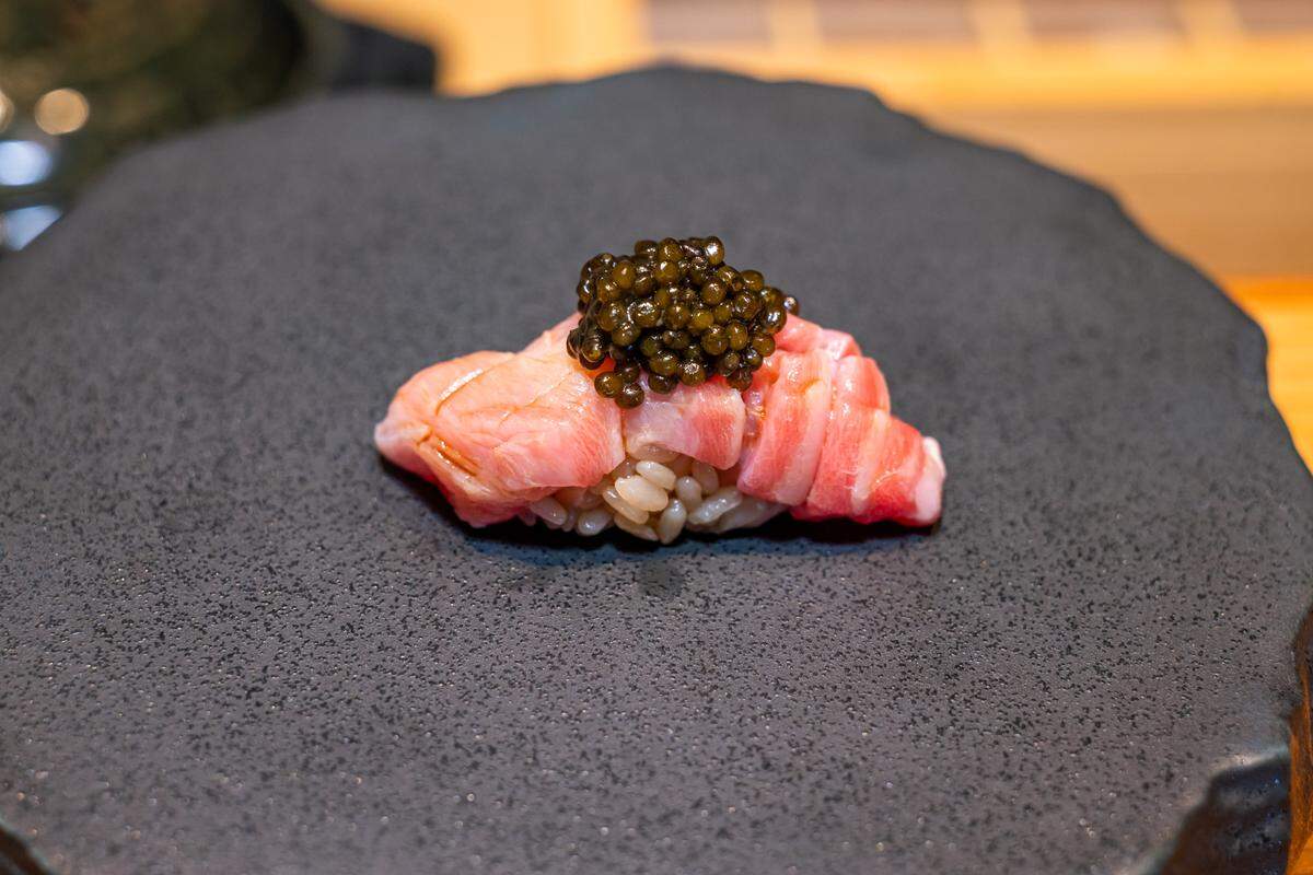 An extreme close-up of a single piece of nigiri sushi topped with a generous portion of dark, glossy caviar, presented on a dark, textured, stone-like plate.