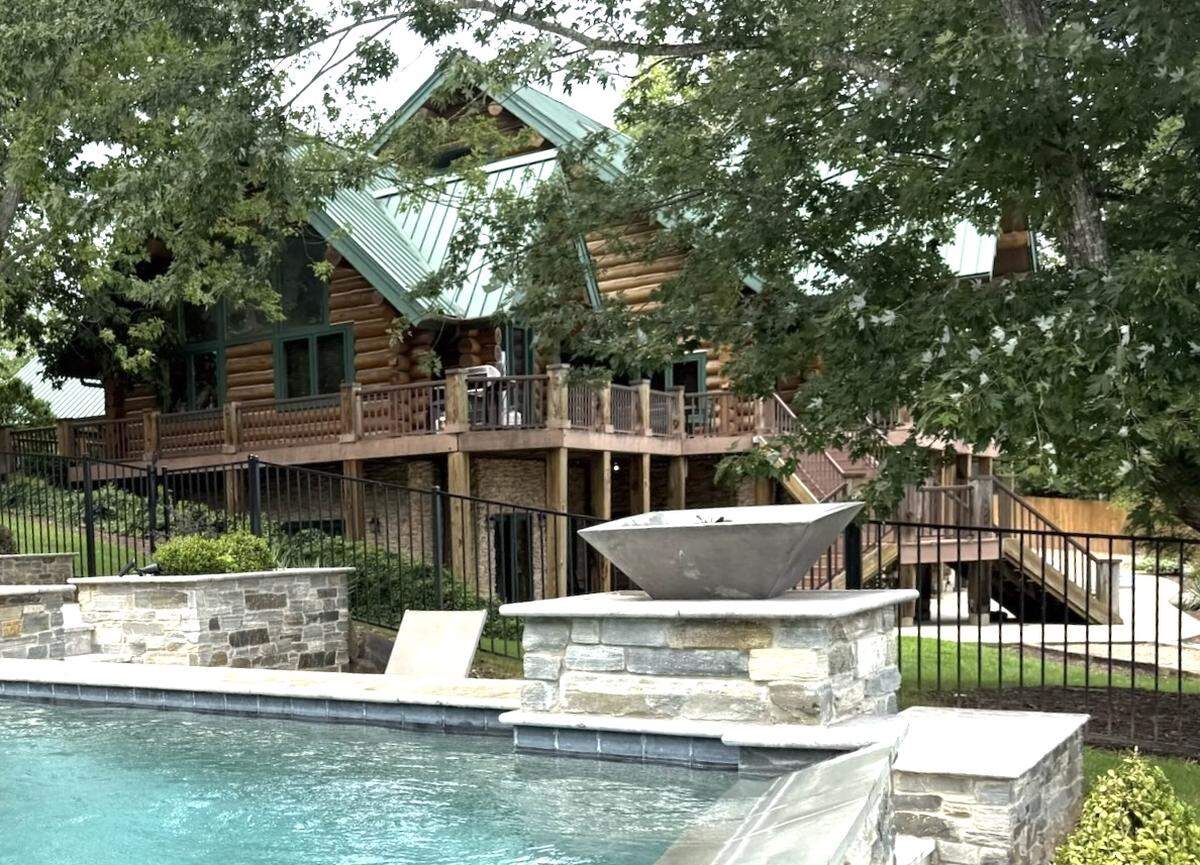 A view from a stone pool deck, looking across the turquoise water of a swimming pool toward a large, multi-level log cabin with a green roof. The cabin sits on a hill behind a black metal fence and is partially obscured by trees.