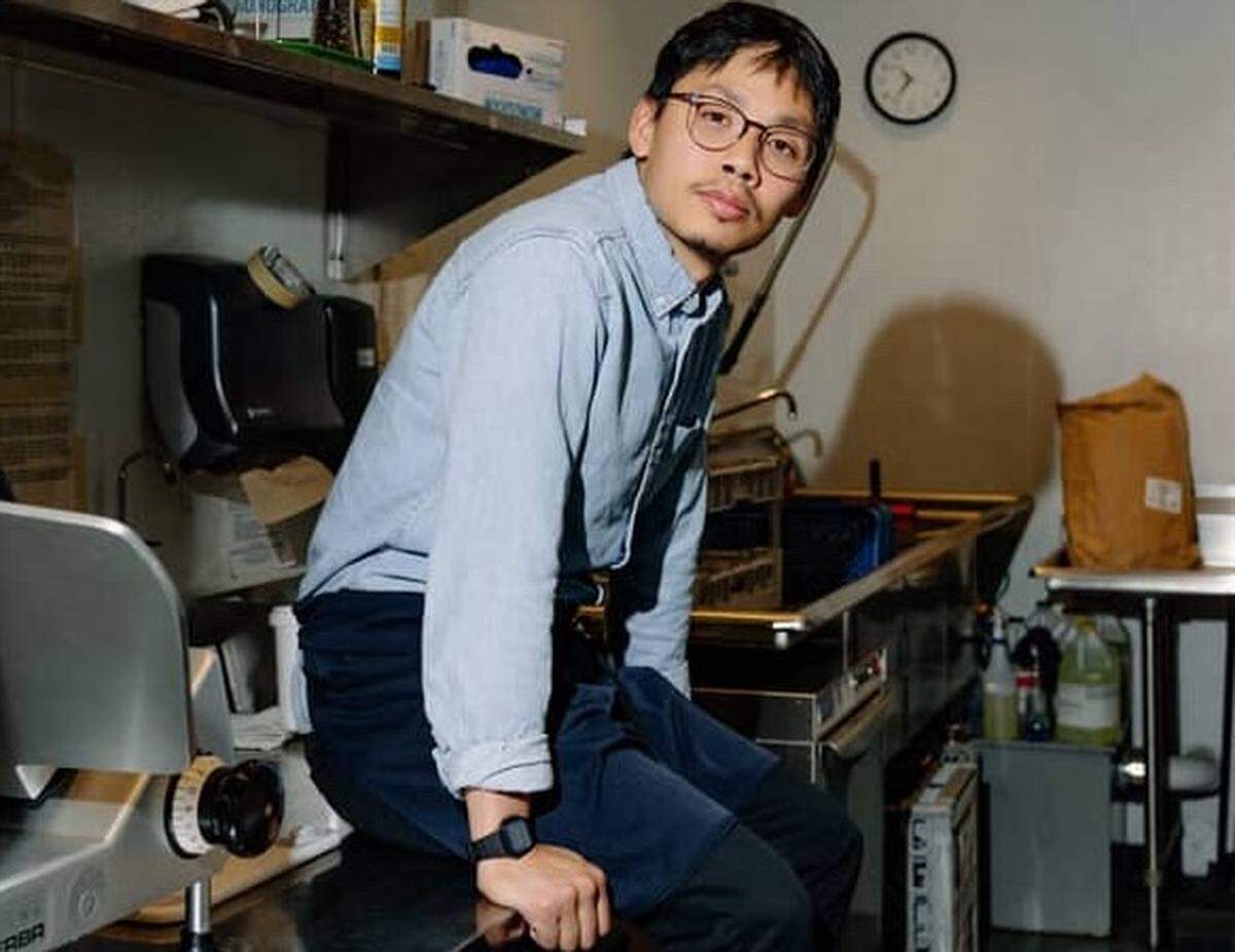 A portrait of a chef wearing a light blue button-down shirt and a navy blue apron, sitting on a stainless steel counter in a commercial kitchen and looking directly at the camera with a neutral expression. The background shows kitchen equipment, including a slicer on the left, a clock on the wall, and various storage containers.