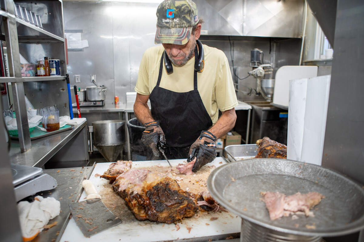 In the kitchen of Red Bridges BBQ, a pitmaster concentrates on carving a large, juicy pork shoulder while standing at a large cutting board with a cleaver nearby. The meat has a dark, seasoned crust, and the pitmaster is separating it into smaller pieces.