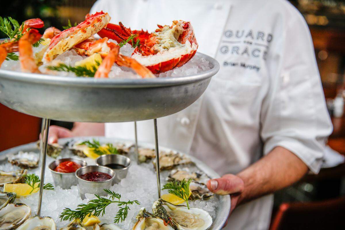 This is a vibrant, close-up photograph of a two-tiered chilled seafood tower being held by a chef at Guard and Grace. The image emphasizes the freshness and luxury of the restaurant’s raw bar offerings.
