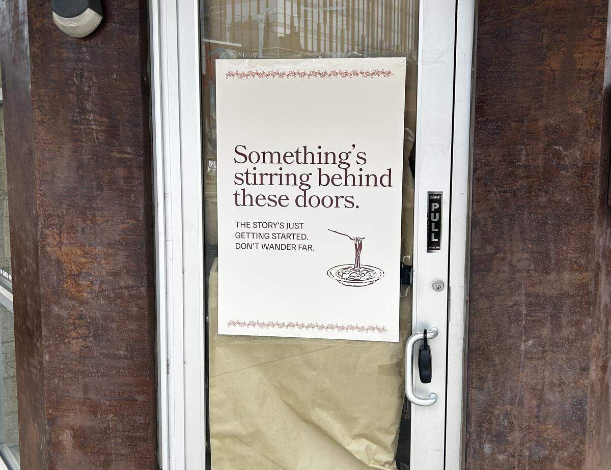 A close-up of a glass storefront door with an opaque interior. A promotional poster taped to the glass features red text that reads: “Something’s stirring behind these doors. THE STORY’S JUST GETTING STARTED. DON’T WANDER FAR.” The poster also includes a small graphic of twirling pasta.