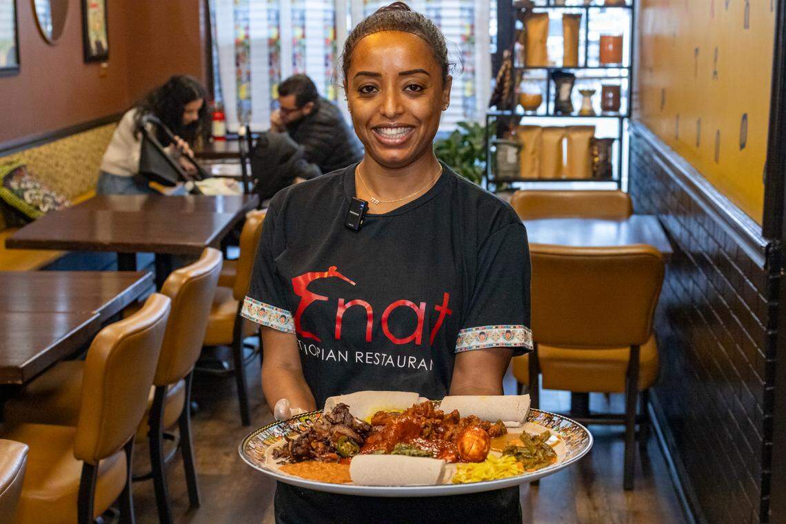 A portrait of a smiling restaurateur in the dining room, proudly holding a large, completed communal platte while wearing a black “Enat” t-shirt and clear serving gloves. In the background, diners can be seen enjoying their meals, creating a sense of community and authentic hospitality.