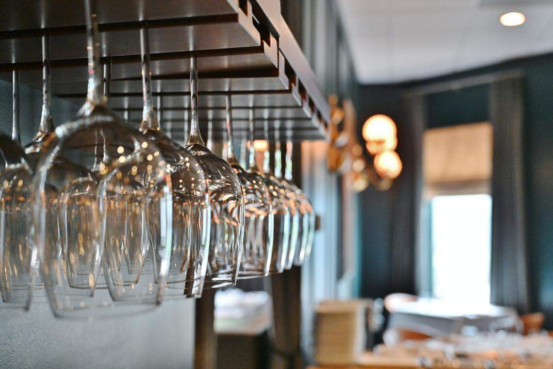 A close-up, shallow-focus shot of several rows of clean wine glasses hanging upside down from a dark wooden overhead rack. The background is softly blurred, showing the warm ambient lighting and elegant interior of a restaurant.