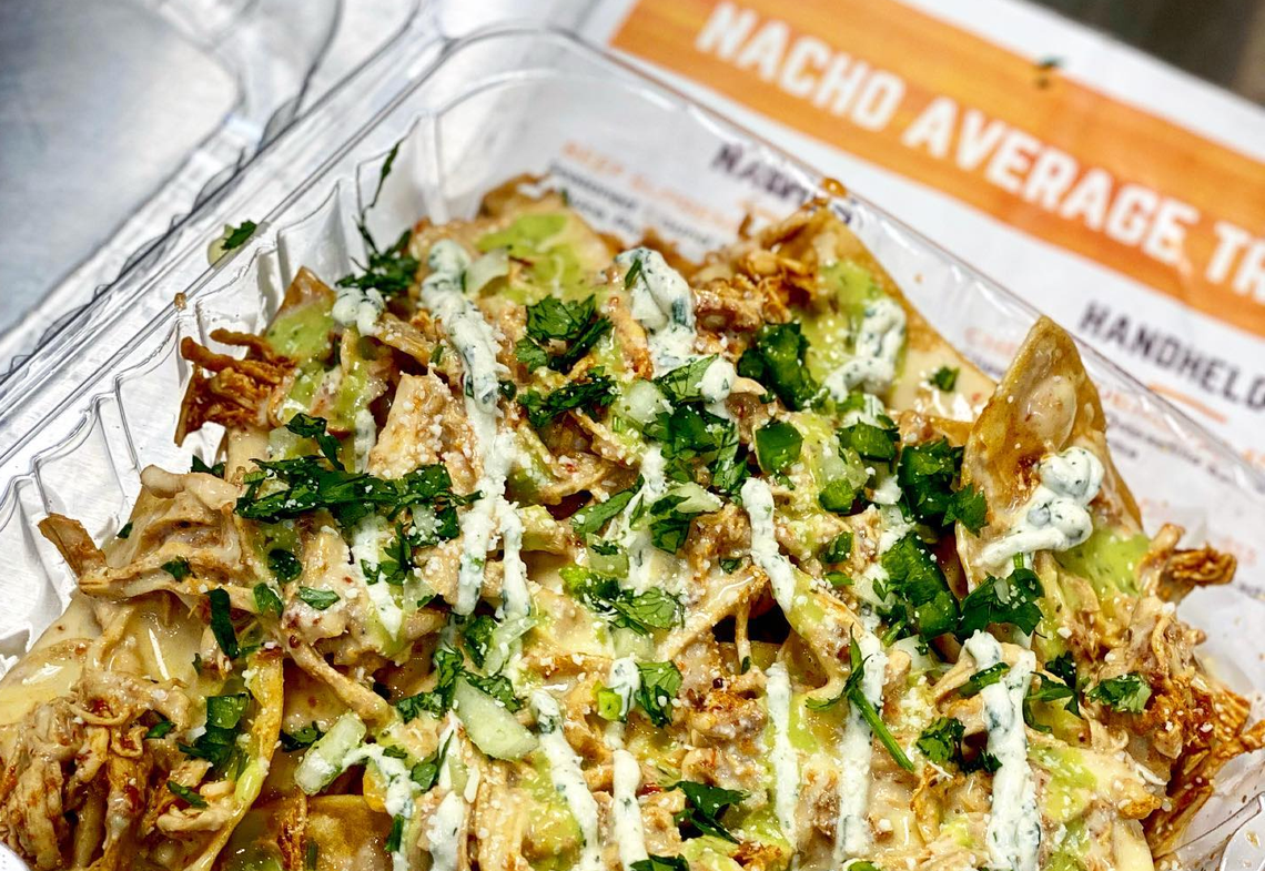 A close-up, high-angle shot of a plastic takeout container filled with a large serving of nachos. The nachos are piled high with crispy tortilla chips, shredded chicken or pork, and a white cheese sauce. They are generously topped with a green creamy sauce, a white drizzle, and chopped fresh cilantro and green onions. In the background, the logo of “NACHO AVERAGE TRUCK” is visible.